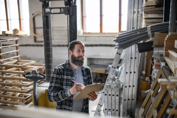 Mature male carpenter doing an inventory indoors in carpentery workshop. Small business concept.