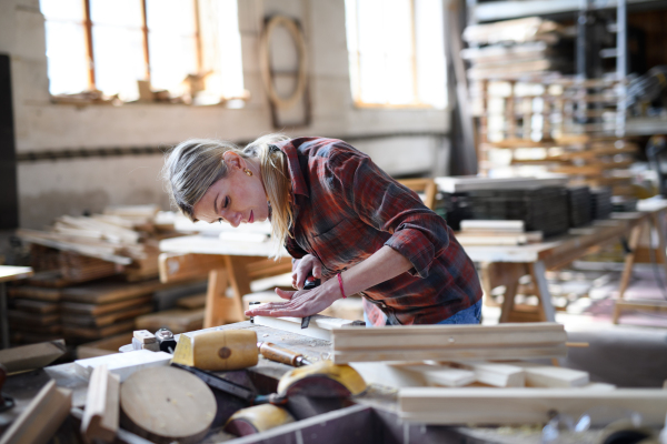 A portrait of female carpenter with goggles working on her product indoors in carpentry workshop. Small business concept.