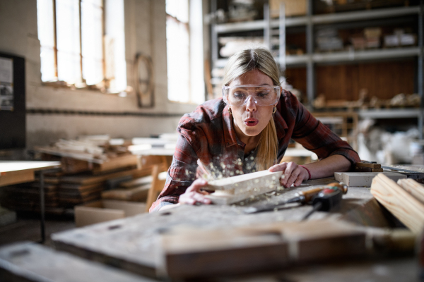 A female carpenter with goggles working on her product indoors in carpentry workshop.