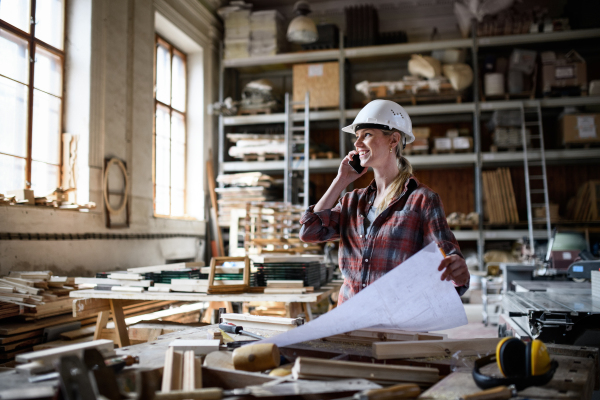 A female engineer holding blueprints and making phone call indoors in carpentry workshop.