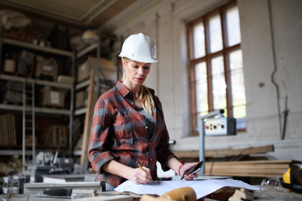 A portrait of female engineer looking at blueprints indoors in carpentry workshop. Small business concept.