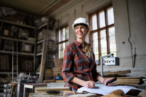 A female engineer holding blueprints and smartphone indoors in carpentry workshop.