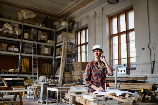 A female engineer holding blueprints and making phone call indoors in carpentry workshop.