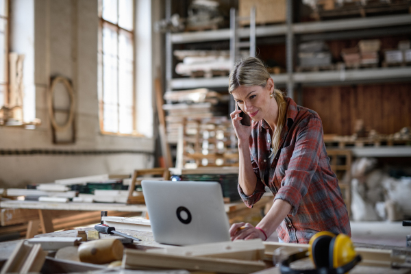 A mid adult woman using laptop and smartphone indoors in carpentry workshop, small business concept.