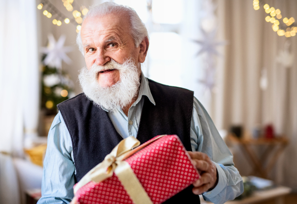 Front view of happy senior man with present box indoors at home at Christmas, looking at camera.