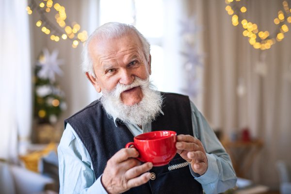 Front view of happy senior man with cup of tea indoors at home at Christmas, looking at camera.