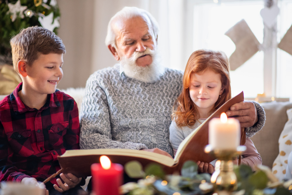 Portrait of small children with senior grandfather indoors at home at Christmas, looking at photographs.