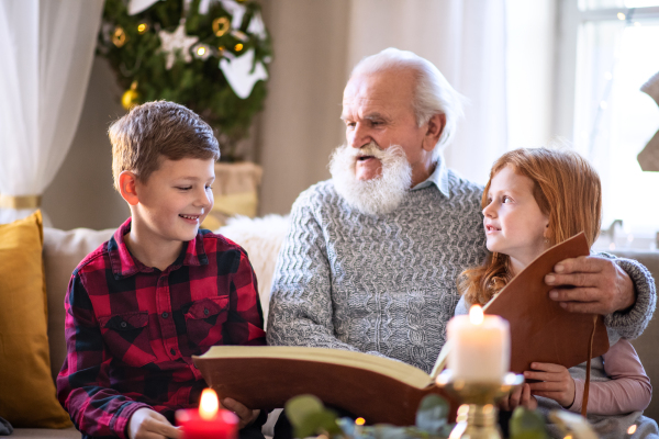 Portrait of small children with senior grandfather indoors at home at Christmas, looking at photographs.