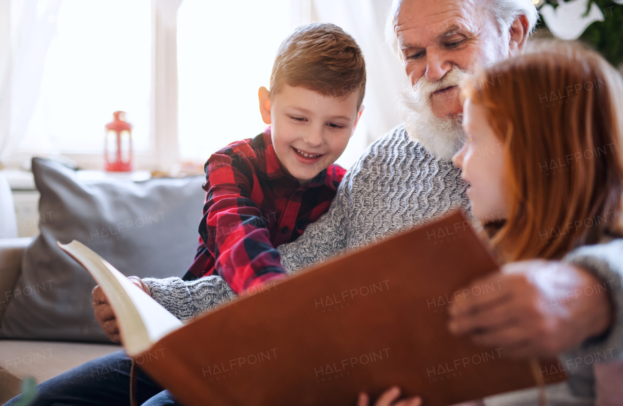 Portrait of small children with senior grandfather indoors at home at Christmas, looking at photographs.