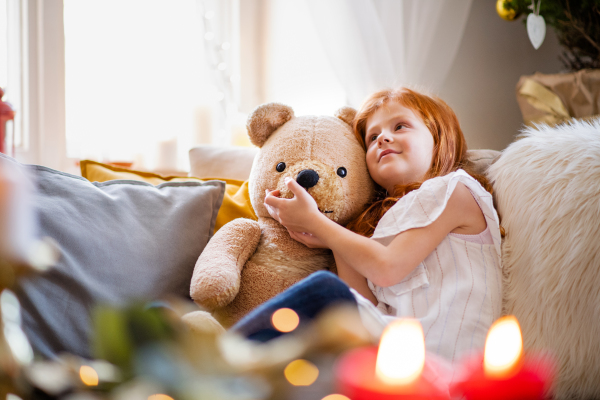 Happy small girl sitting on sofa indoors at home at Christmas, hugging teddy bear.