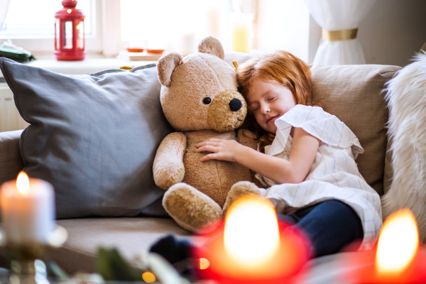 Small girl lying and sleeping on sofa indoors at home at Christmas, hugging teddy bear.