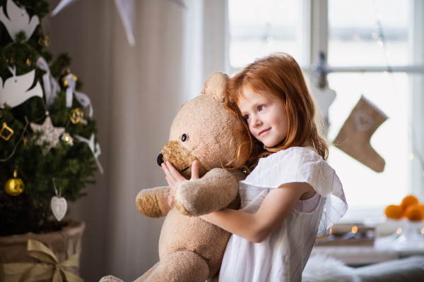 Portrait of small girl standing indoors at home at Christmas, hugging teddy bear.