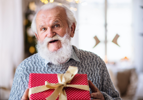 Front view of happy senior man with present box indoors at home at Christmas, looking at camera.