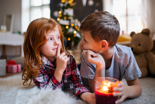 Portrait of small girl and boy with candle indoors at home at Christmas, making silence gesture.