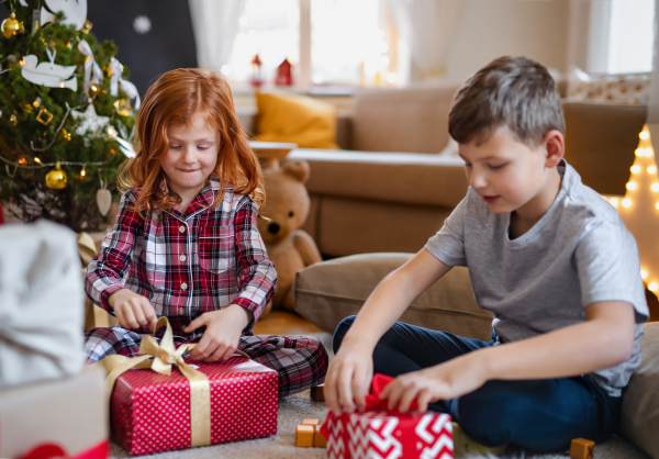 Small girl and boy in pajamas indoors at home at Christmas, opening presents in the morning.