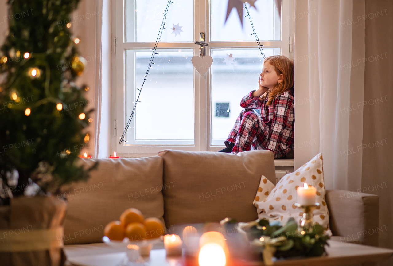 Side view of portrait of small sad girl in pajamas indoors at home at Christmas, sitting on window.