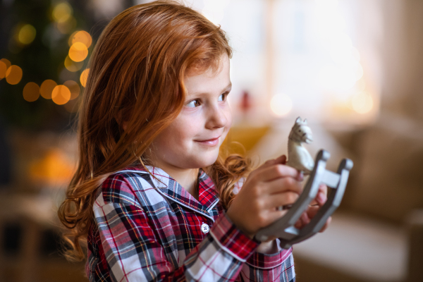 Side view portrait of small girl indoors at home at Christmas, holding wooden horse.