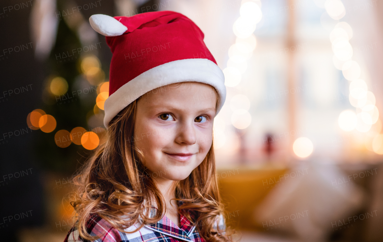 Close-up portrait of small girl with Santa hat indoors at home at Christmas, looking a camera.