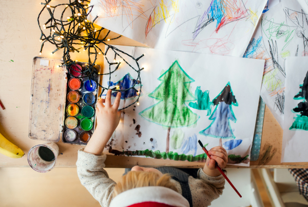 Top view of unrecognizable small child indoors at home at Christmas, painting pictures with waterpaints.