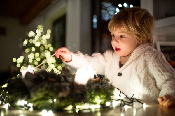 Portrait of small boy indoors at home at Christmas, lightning candles on wreath.