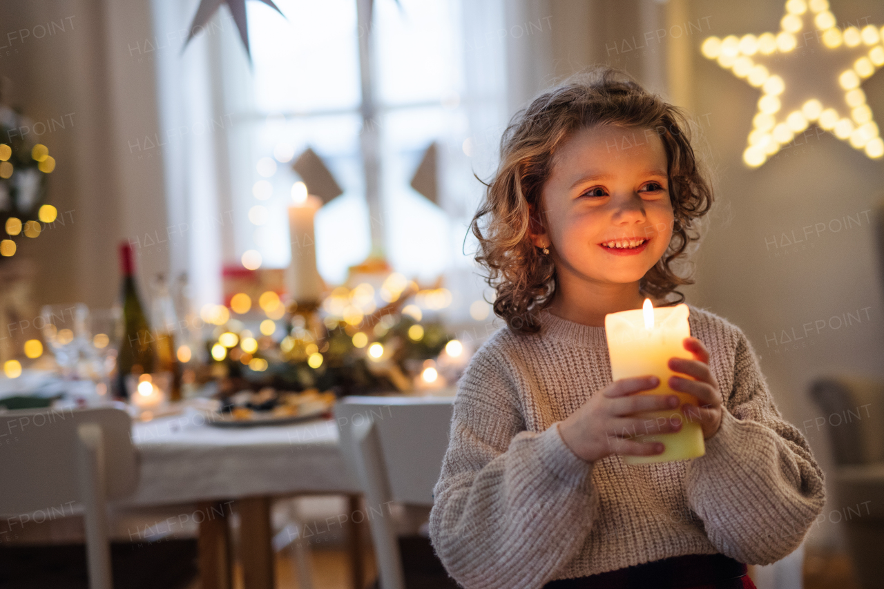 Front view of cheerful small girl standing indoors at Christmas, holding candle.