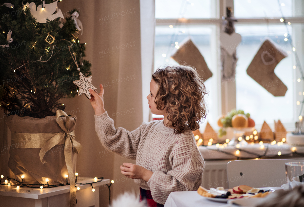 Side view of happy small girl indoors at home, standing by Christmas tree.