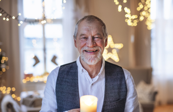 A front view of happy senior man indoors holding candle at Christmas.