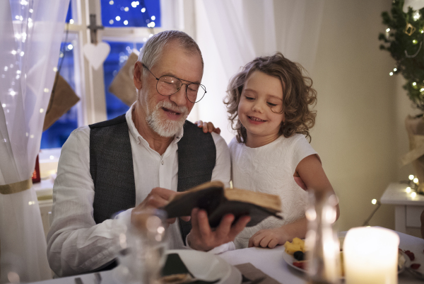 Happy senior grandfather with small granddaughter indoors at Christmas, reading Bible book.