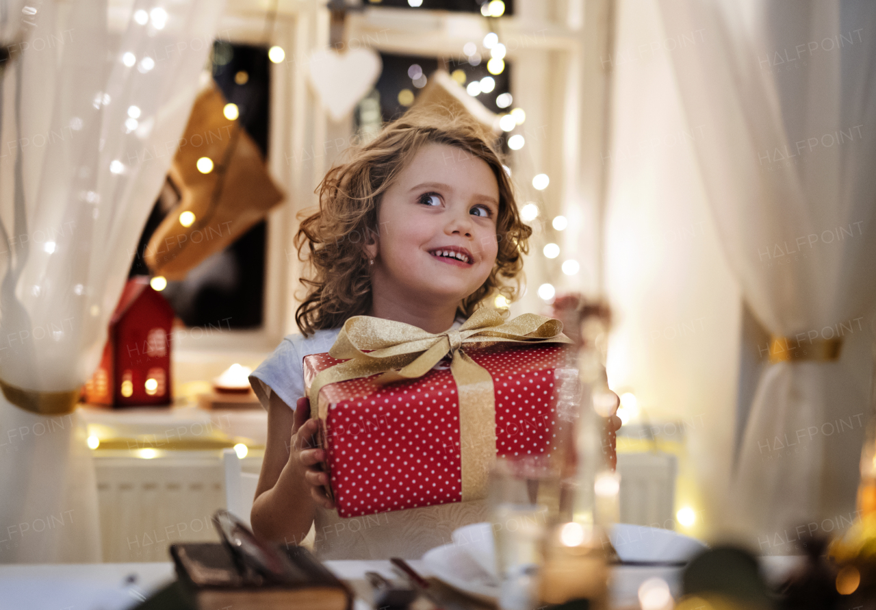 Front view of cheerful small girl sitting indoors at Christmas, holding present.