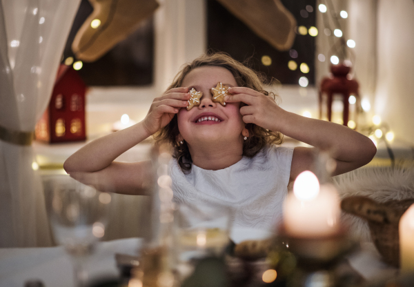 Cheerful small girl sitting indoors at Christmas, covering eyes with gingerbread biscuit.