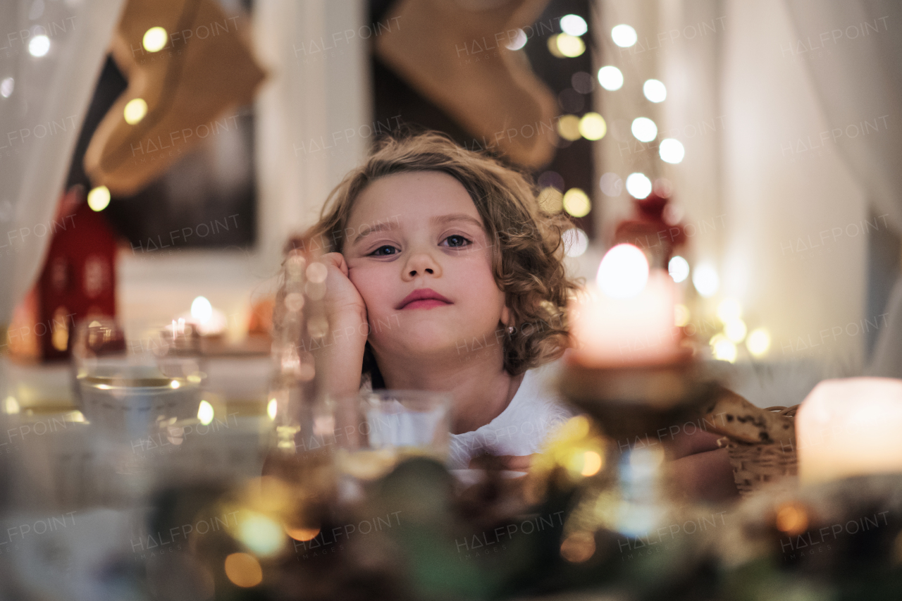 Portrait of bored small girl sitting at the table indoors at Christmas.