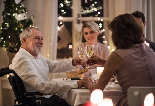 Senior man in wheelchair with family indoors celebrating Christmas together, clinking glasses.