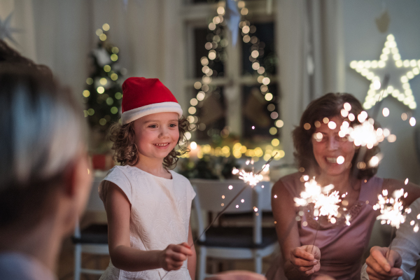 Small girl with parents and grandparents indoors celebrating Christmas, holding sparklers.