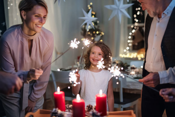 Small girl with parents and grandparents indoors celebrating Christmas, holding sparklers.