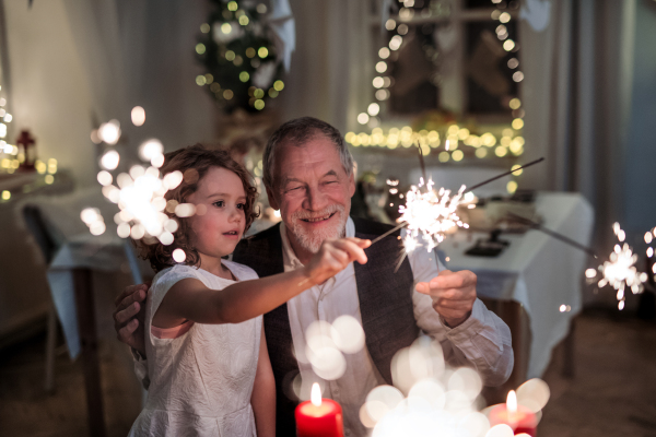 Portrait of senior grandfather with small granddaughter indoors at Christmas, having fun with sparklers.