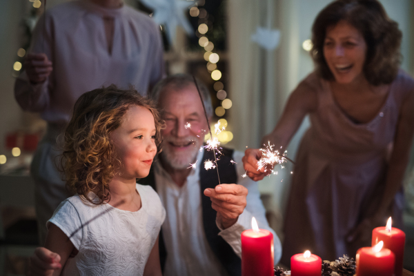 Small girl with parents and grandparents indoors celebrating Christmas, holding sparklers.