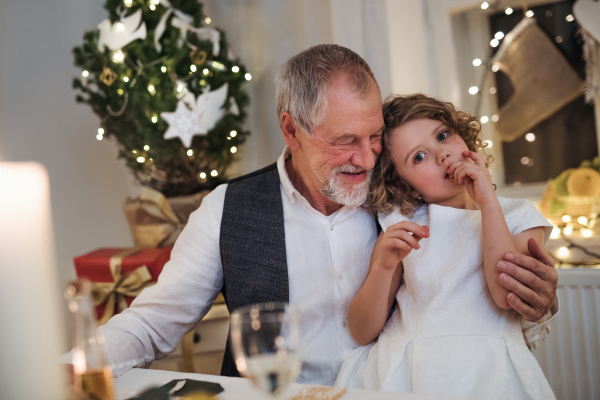 Happy senior grandfather with small granddaughter indoors at Christmas, sitting at table.