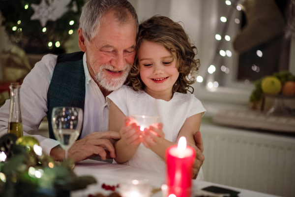 Happy senior grandfather with small granddaughter indoors at Christmas, sitting at table.