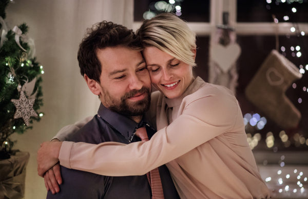 Front view of happy couple sitting indoors at the table celebrating Christmas, hugging.