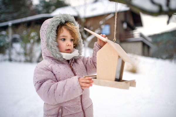 Portrait of small girl outdoors in winter garden, standing by wooden bird feeder.