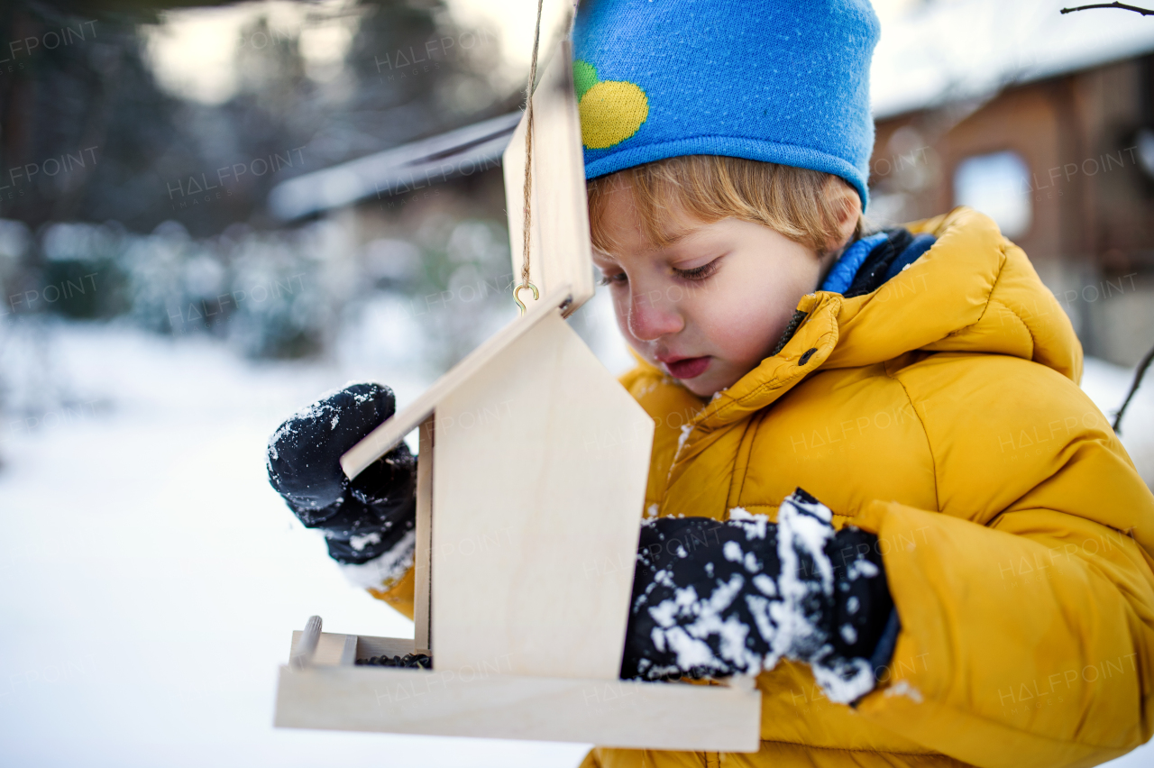 Portrait of small girl outdoors in winter garden, standing by wooden bird feeder.