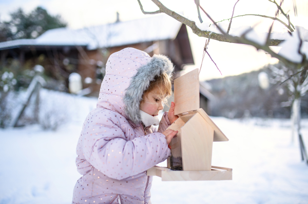 Portrait of small girl outdoors in winter garden, standing by wooden bird feeder.