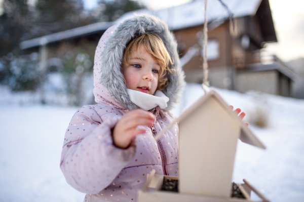 Portrait of small girl outdoors in winter garden, standing by wooden bird feeder.