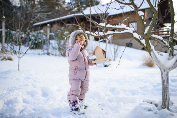 Portrait of small girl outdoors in winter garden, standing by wooden bird feeder.