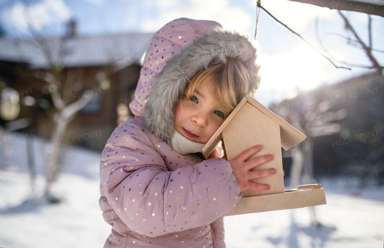 Portrait of small girl outdoors in winter garden, standing by wooden bird feeder.