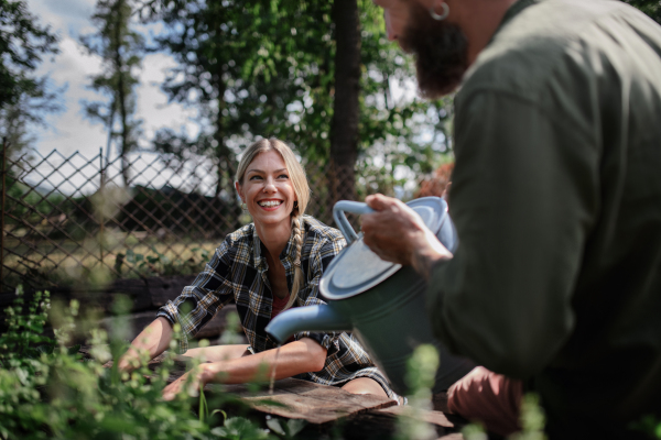 A happy mid woman farmer looking at unrecognizable man watering plants outdoors in garden.