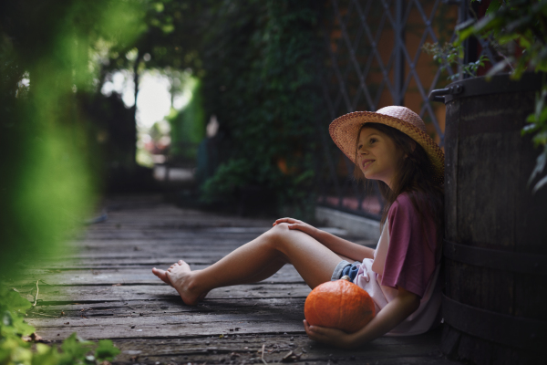 A little girl in hat sitting and holding organic pumpkin outdoors at farm.