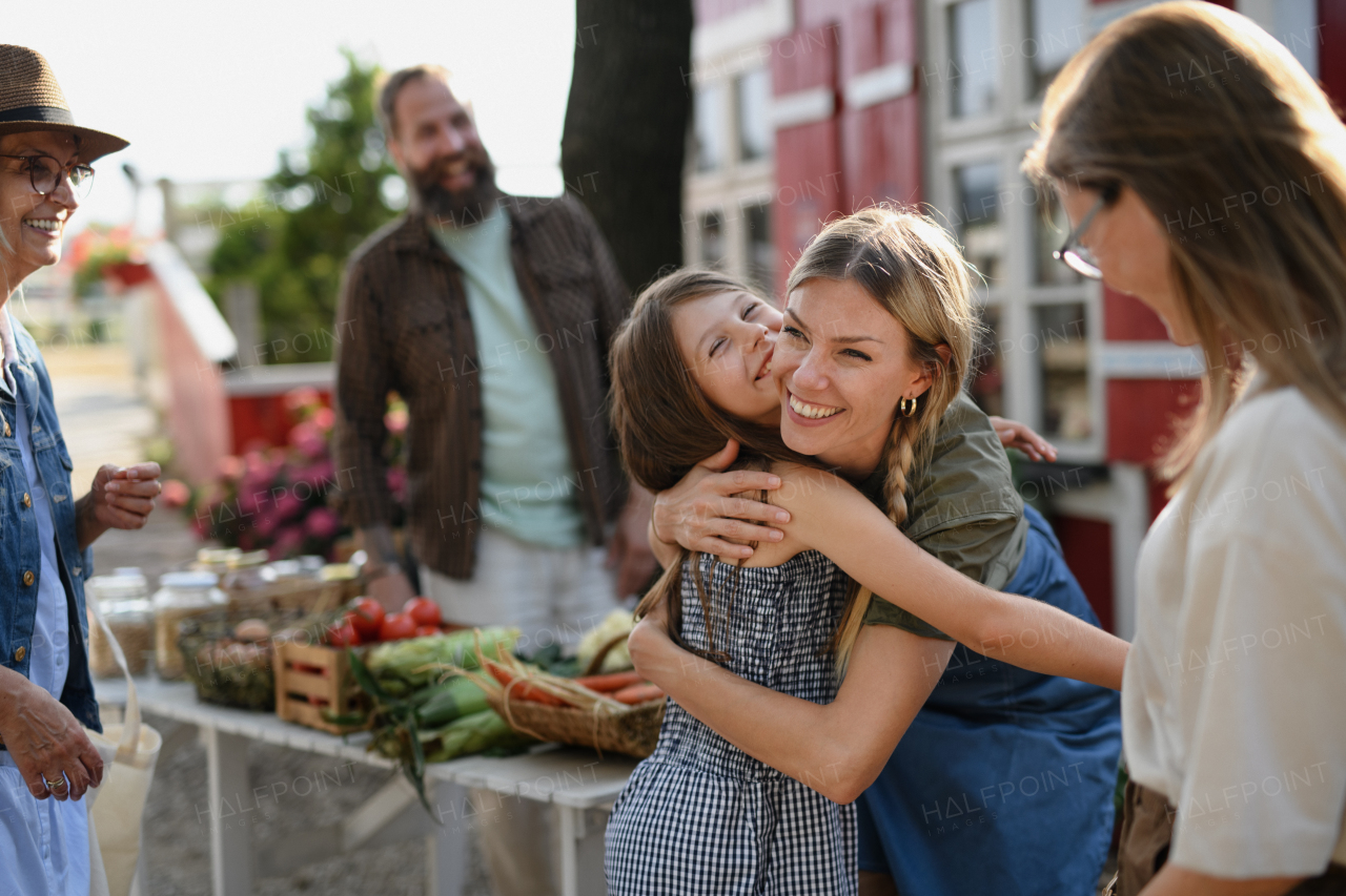 A little girl with mother hugging outdoors at community farmers market.