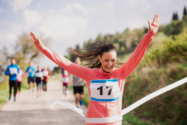 A young woman runner crossing finish line in a race competition in nature, arms raised.