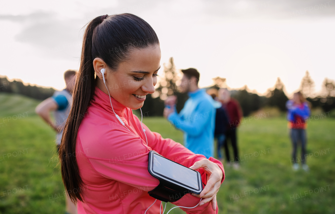 A portrait of young woman with earphones and group of people doing exercise in nature, resting.